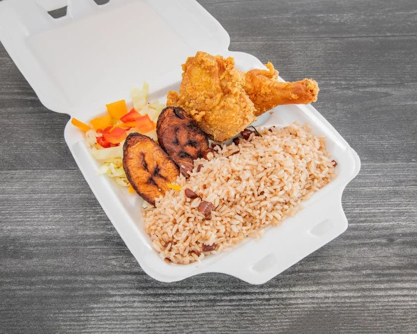 Crispy fried chicken with rice and peas, fried plantains, and cabbage medley in a white takeout container