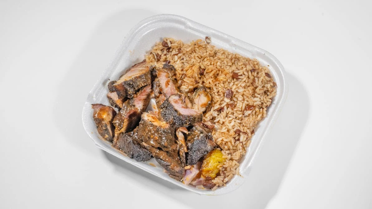 Takeout container of chopped Jamaican jerk pork glazed in dark savory sauce with rice and peas and fried plantain